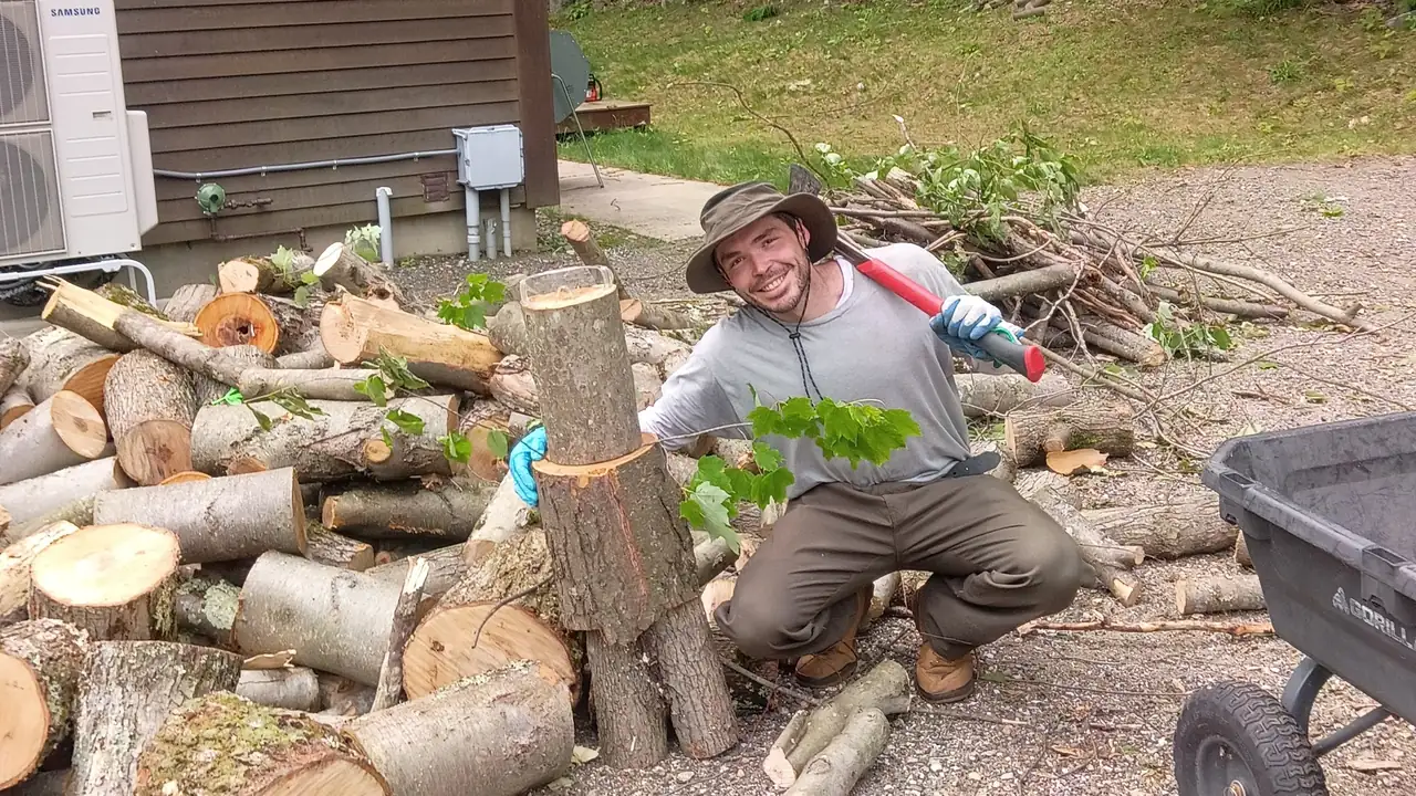 JMR owner holding a splitting maul beside a pile of cut logs during a yard cleanup